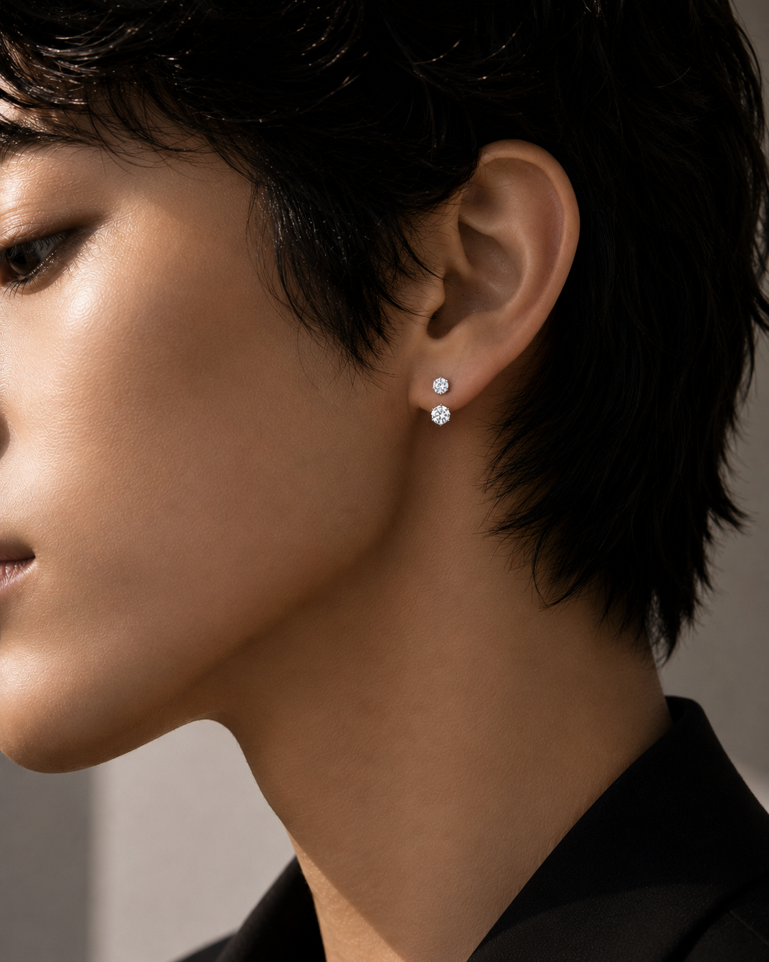 Floating double diamond earrings worn on an androgynous model, featuring a concealed rear floating structure with no visible front connection. The upper round brilliant diamond sits at the ear piercing, while the slightly larger lower diamond rests just beneath the earlobe edge, creating a subtle suspended effect. Editorial close-up jewelry campaign image with strong diamond fire, minimal styling, and modern architectural luxury aesthetic.