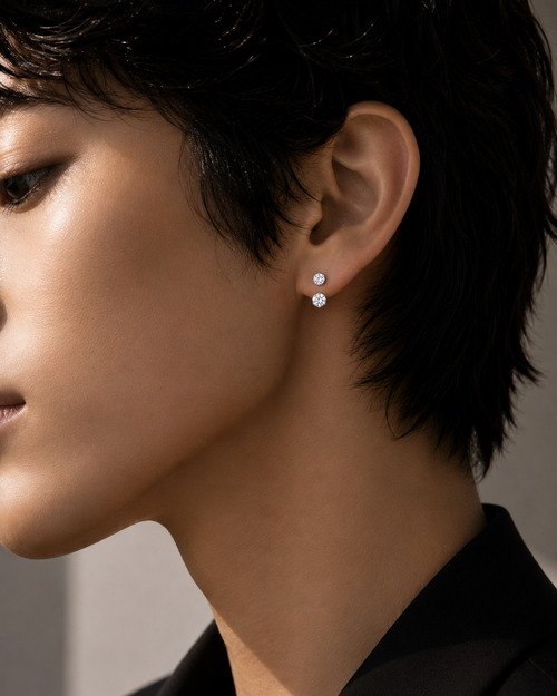 Floating double diamond earrings worn on an androgynous model, featuring a concealed rear floating structure with no visible front connection. The upper round brilliant diamond sits at the ear piercing, while the slightly larger lower diamond rests just beneath the earlobe edge, creating a subtle suspended effect. Editorial close-up jewelry campaign image with strong diamond fire, minimal styling, and modern architectural luxury aesthetic.